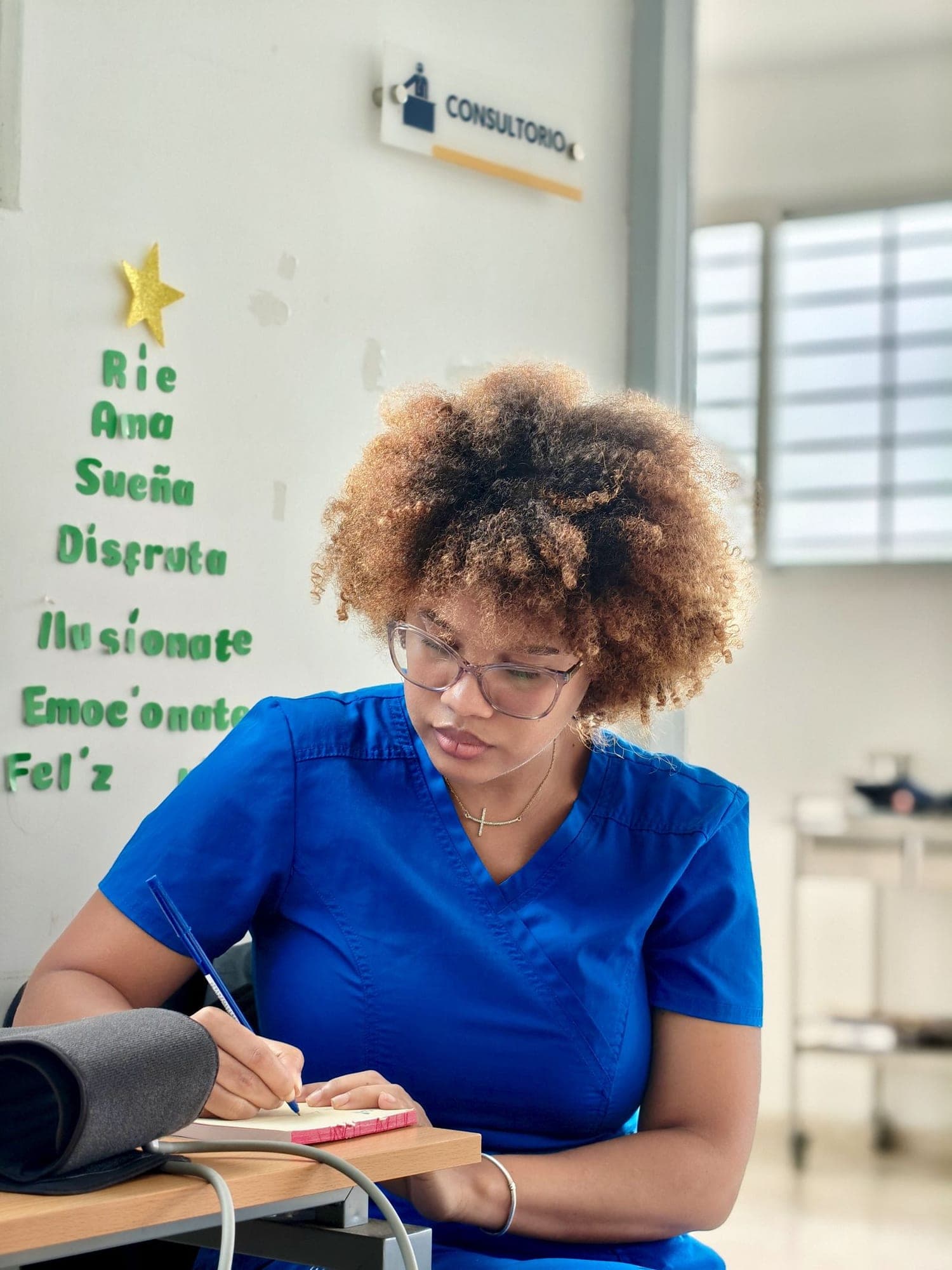 Médica con uniforme azul escribe notas en la mesa de su consultorio, junto al tensiómetro